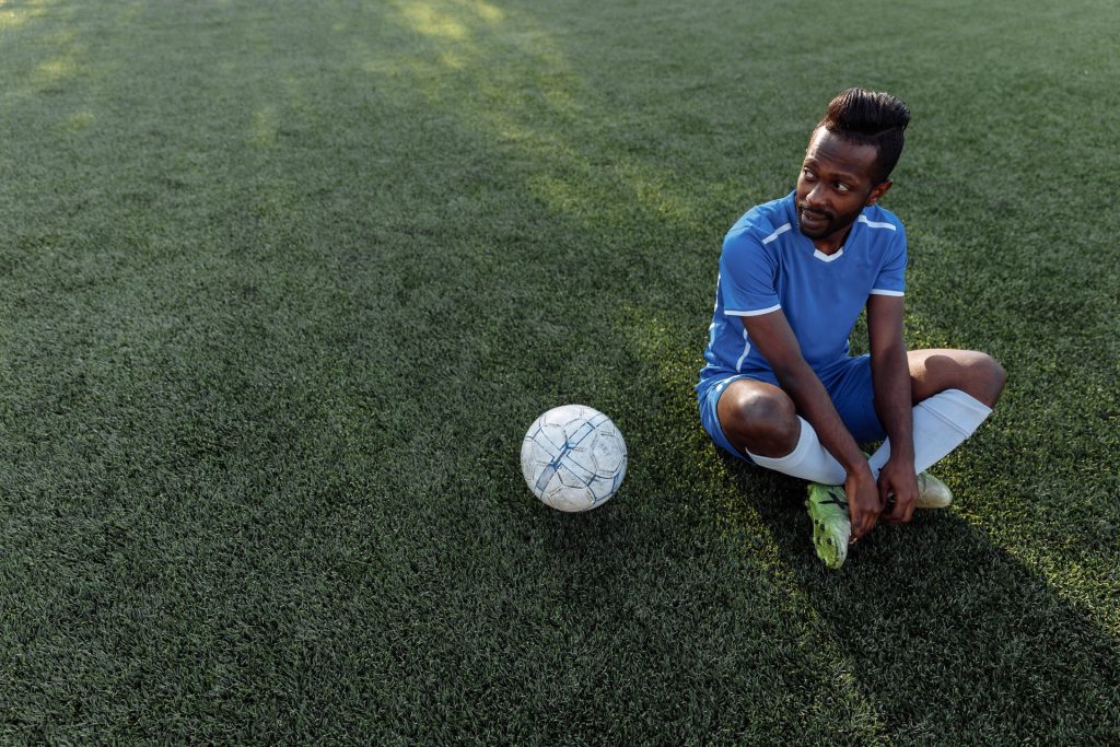 A footballer sitting on the pitch beside the ball during a break in play