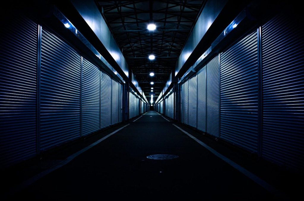 Long corridor lined with closed metal roller shutters under ceiling lights