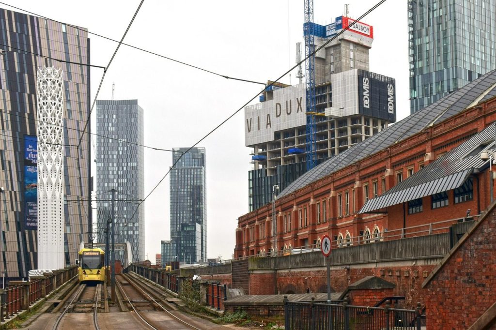 Red brick former mill building beside a Manchester tram line with new high-rise towers behind